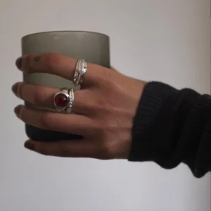 Hand holding frosted glass cup, wearing sterling silver rings including a January Birthstone Ring with natural garnet in teardrop bezel, styled with textured bands and dark sleeve.