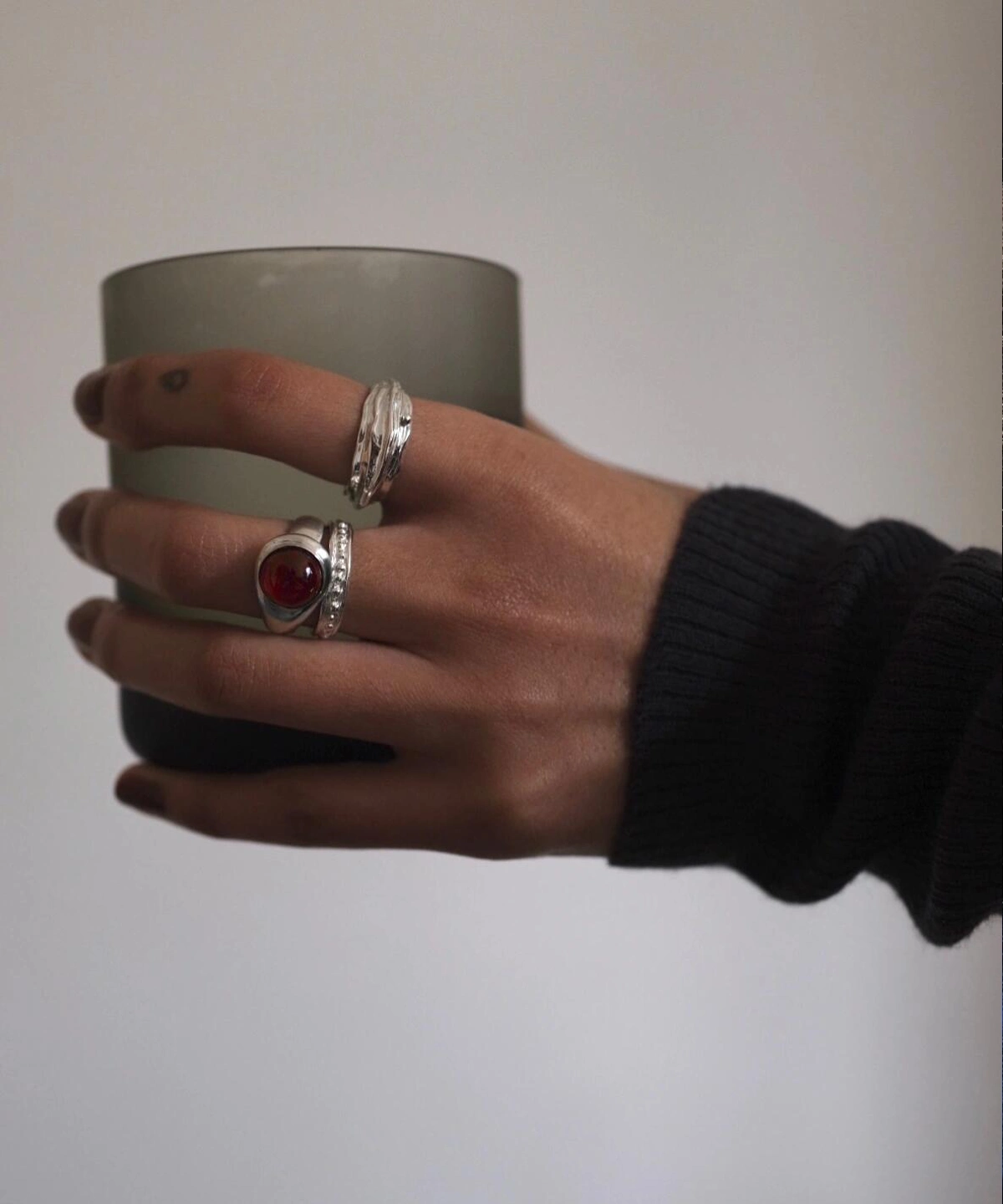 Hand holding frosted glass cup, wearing sterling silver rings including a January Birthstone Ring with natural garnet in teardrop bezel, styled with textured bands and dark sleeve.