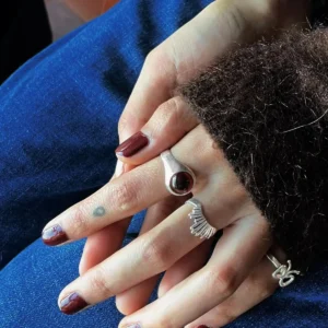 Hands with dark red nails wearing sterling silver rings, including a January Birthstone Ring with natural garnet, styled with heart and fan motifs over denim and corduroy.