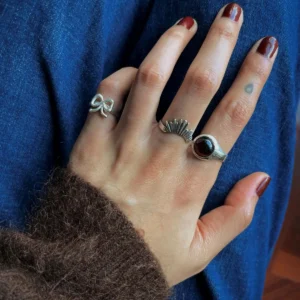 Hand resting on denim wearing sterling silver rings, including a January Birthstone Ring with natural garnet, styled with bow and fan motifs and a blue-green dot tattoo.
