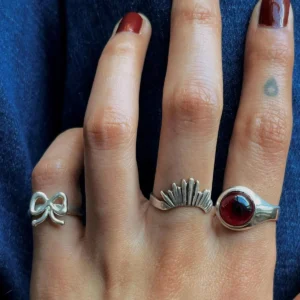 Hand with dark red nails wearing sterling silver rings, including a January Birthstone Ring with natural garnet, styled with bow and fan motifs against dark denim.