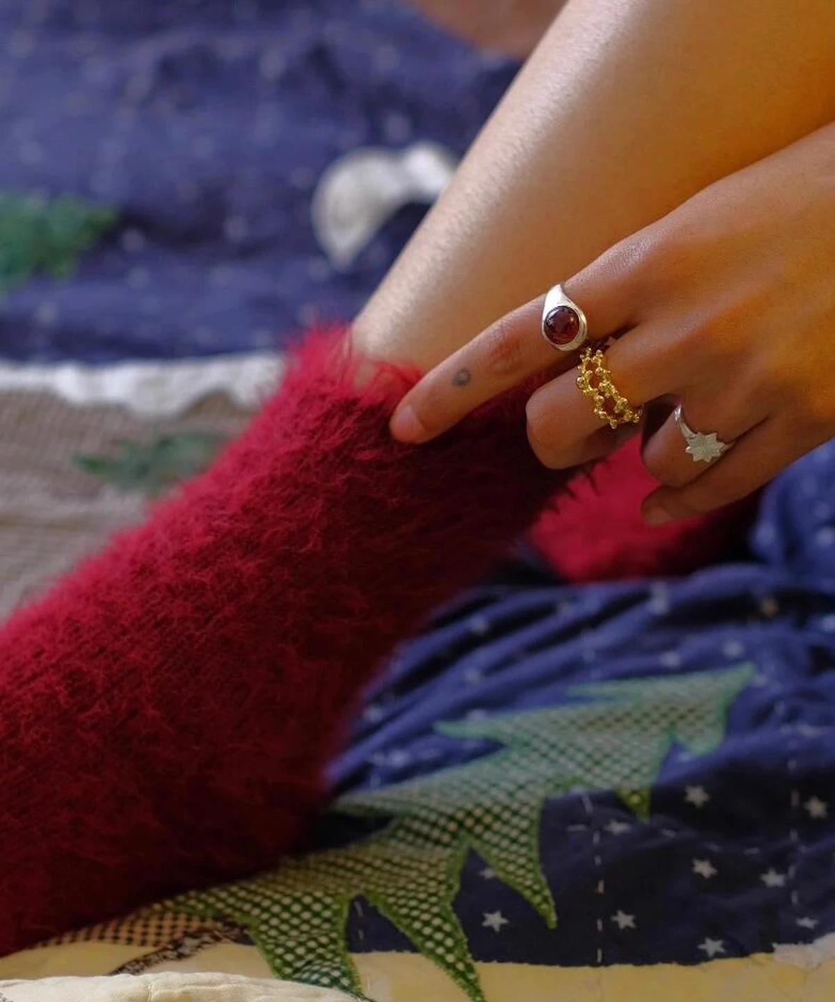 Hand with fuzzy red sock and silver rings, including a January Birthstone Ring with natural garnet, styled with gold and star-shaped accents over a patterned quilt.
