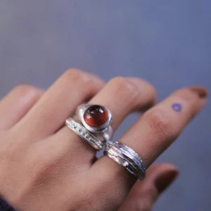 Hand with dark red nails wearing three sterling silver rings, including a January Birthstone Ring with natural garnet, styled with leaf and textured bands against a blurred background.
