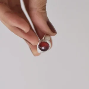 Hand holding a silver ring with a polished red cabochon garnet, styled with dark nails and a light background to highlight the January Birthstone Ring in sterling silver.