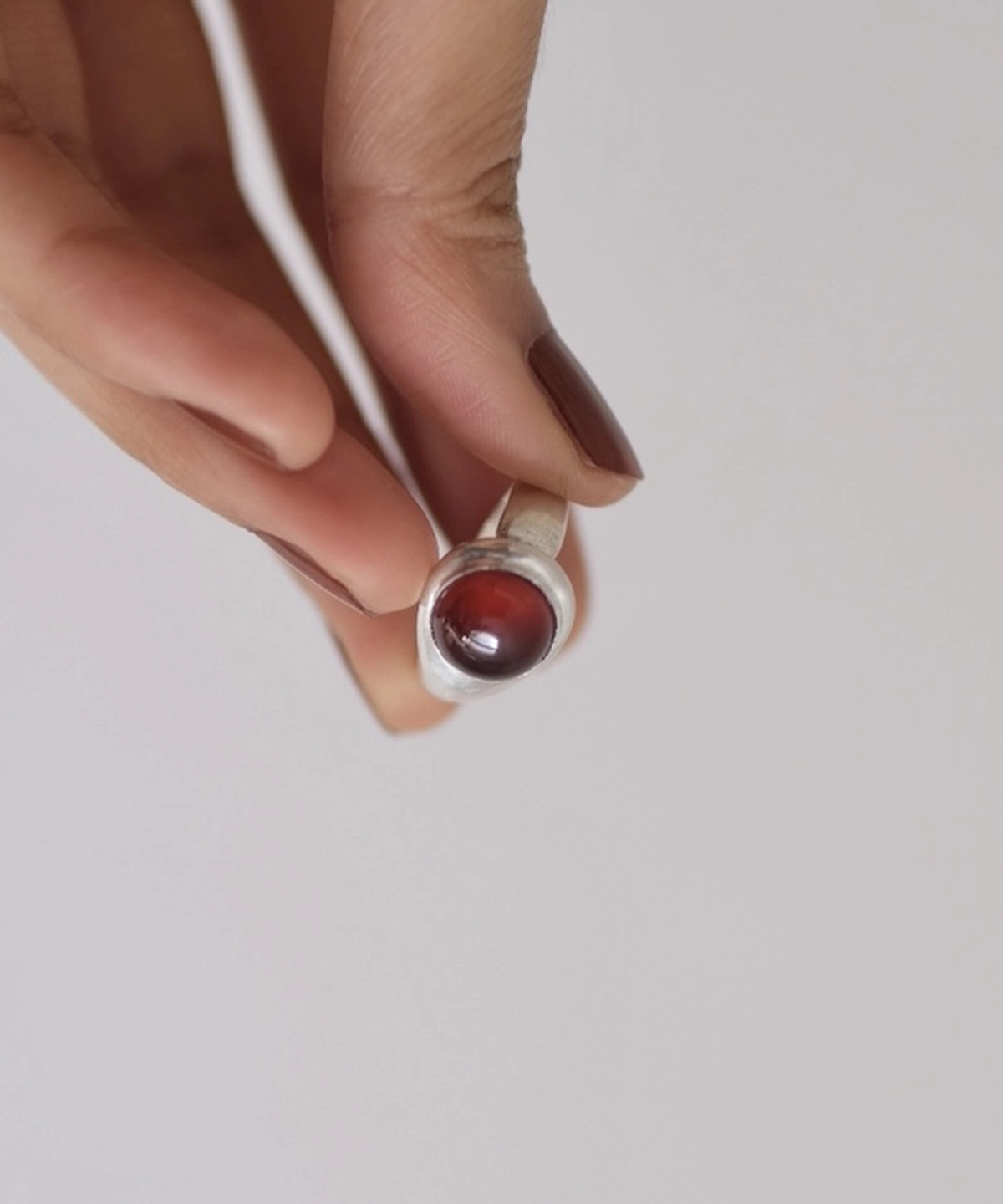 Hand holding a silver ring with a polished red cabochon garnet, styled with dark nails and a light background to highlight the January Birthstone Ring in sterling silver.
