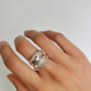 Hand wearing two silver rings on the middle finger, including a July Birthstone Ring with a small natural ruby gemstone and a wavy silver band, styled against a white background.