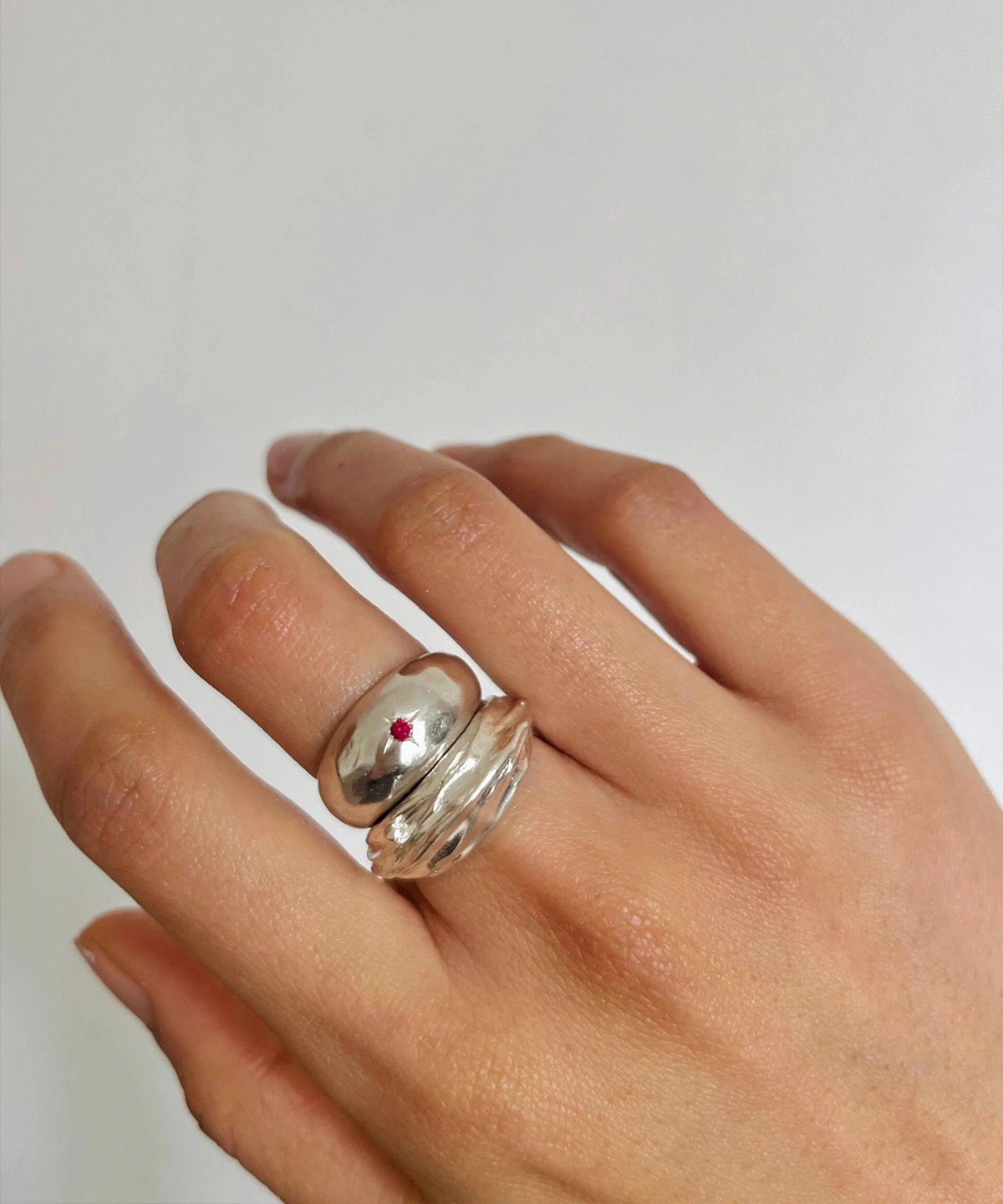 Hand wearing two silver rings on the middle finger, including a July Birthstone Ring with a small natural ruby gemstone and a wavy silver band, styled against a white background.
