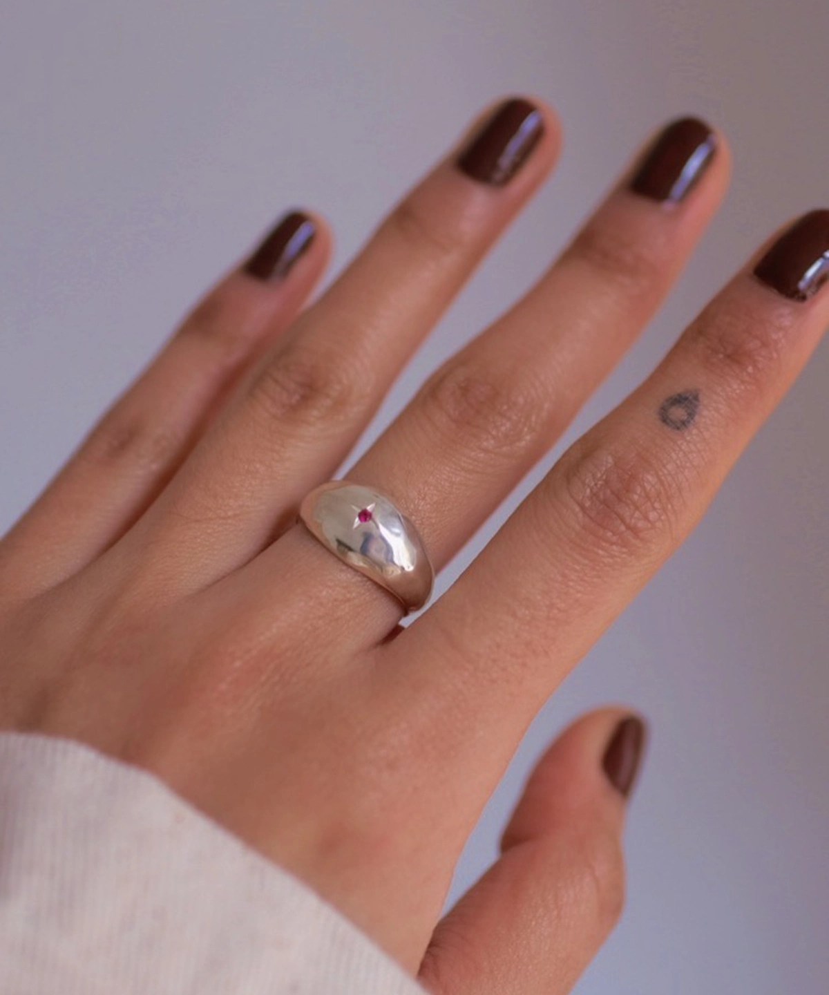 Hand with brown nails wearing a silver ring with a small natural ruby gemstone, styled with a teardrop tattoo and soft sleeve to showcase the July Birthstone Ring’s subtle charm.