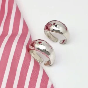 Two open-ended silver rings with hammered texture and small natural ruby gemstones, placed on a white surface beside pink-striped fabric, showcasing the July Birthstone Ring’s artisanal charm.