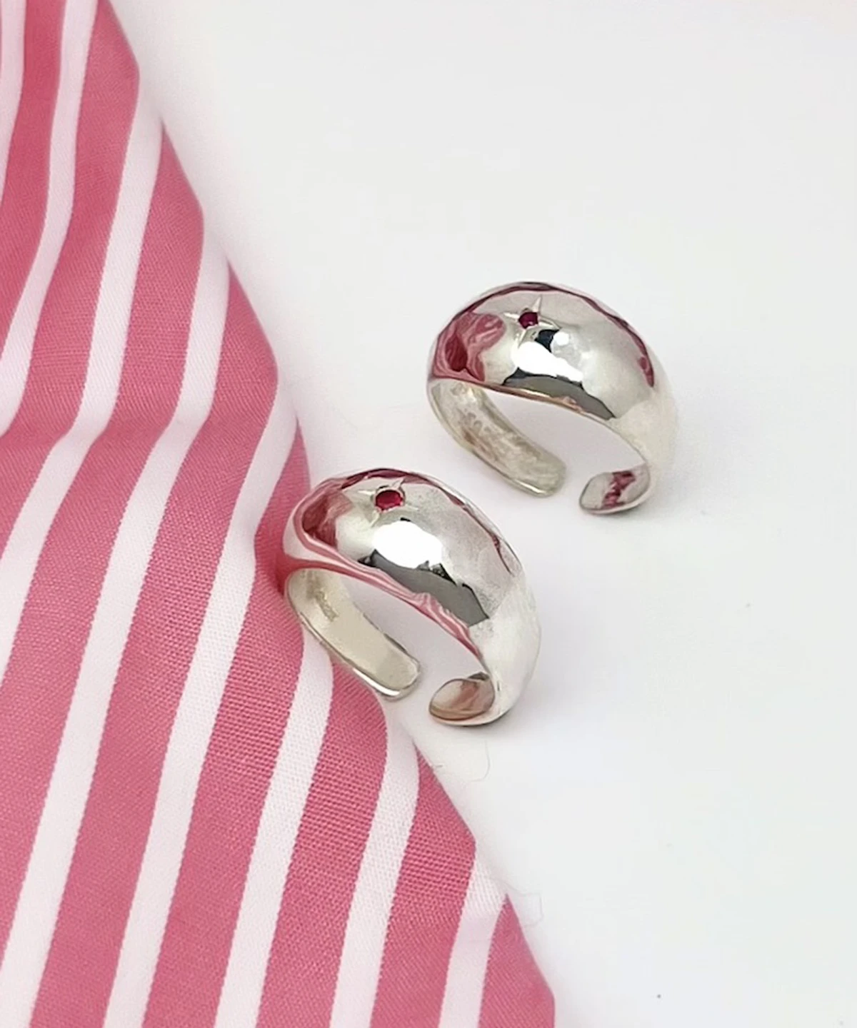 Two open-ended silver rings with hammered texture and small natural ruby gemstones, placed on a white surface beside pink-striped fabric, showcasing the July Birthstone Ring’s artisanal charm.