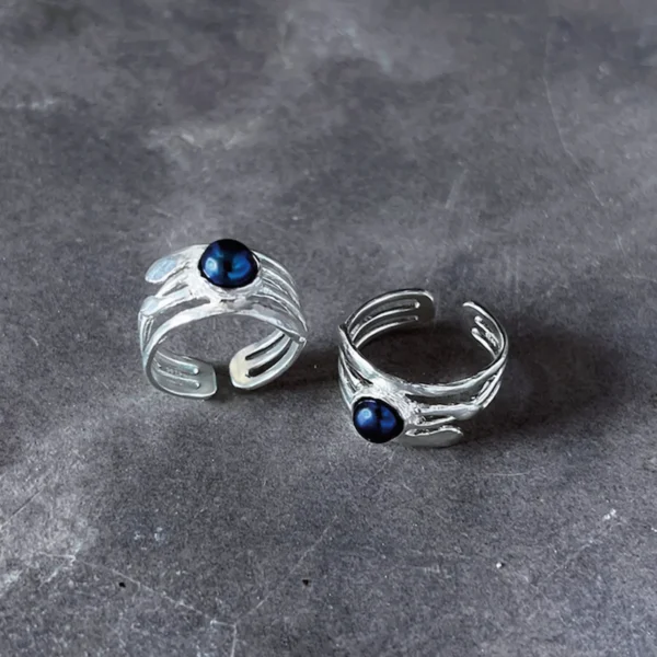 June Birthstone Ring-Natural Black Pearl Ring in Sterling Silver 02 Two handcrafted Silver Rings with natural black pearls displayed on a dark textured surface; elegant June Birthstone Ring designs with flowing metalwork and open bands, showcasing artisanal Black Pearl Ring craftsmanship and poetic styling.