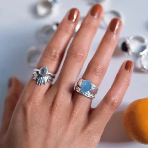 Hand with brown nails wearing silver rings, including a March Birthstone Ring with raw natural aquamarine and teardrop silver detailing, styled beside scattered rings on a white surface.