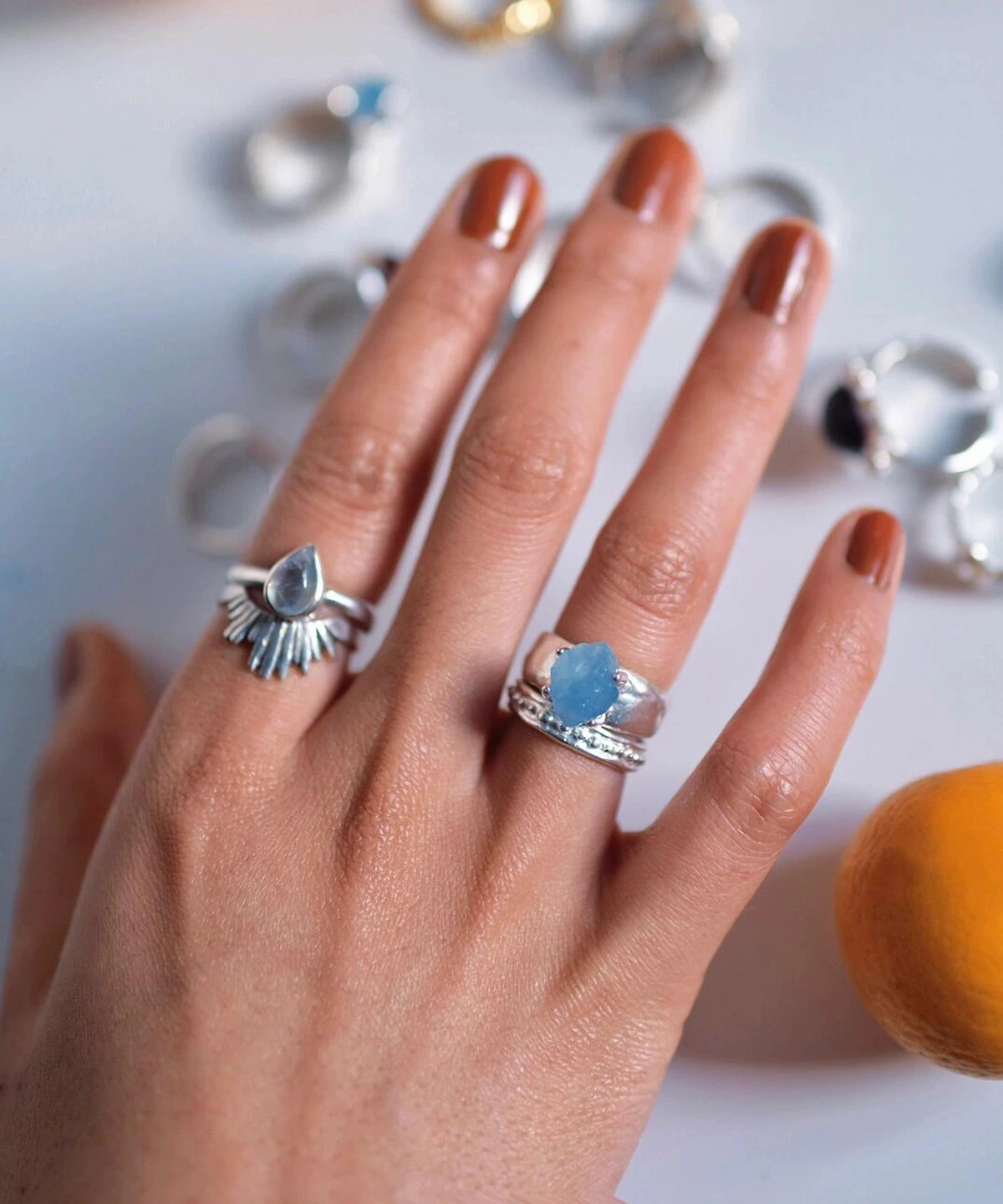 March Birthstone Ring-Natural Aquamarine Ring in Sterling Silver 01 Hand with brown nails wearing silver rings, including a March Birthstone Ring with raw natural aquamarine and teardrop silver detailing, styled beside scattered rings on a white surface.
