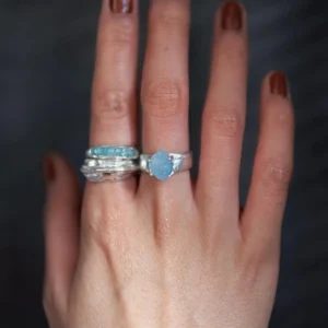 Hand with brown nails wearing silver rings, including a March Birthstone Ring with oval natural aquamarine and stacked bands, styled against a dark blurred background.