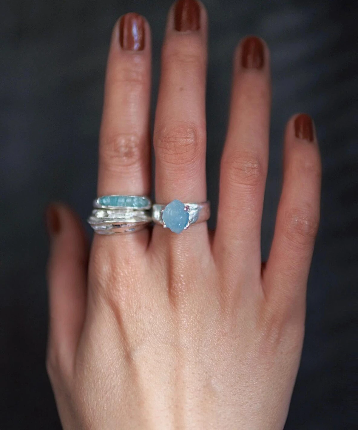 March Birthstone Ring-Natural Aquamarine Ring in Sterling Silver 02 Hand with brown nails wearing silver rings, including a March Birthstone Ring with oval natural aquamarine and stacked bands, styled against a dark blurred background.