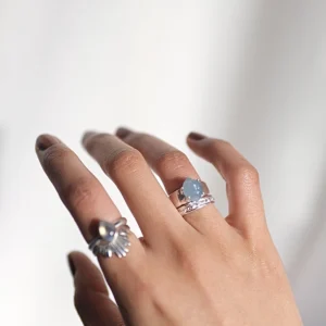 March Birthstone Ring-Natural Aquamarine Ring in Sterling Silver 03 Hand with brown nails wearing silver rings, including a March Birthstone Ring with oval natural aquamarine and a teardrop sunburst design, styled against a soft blurred background.