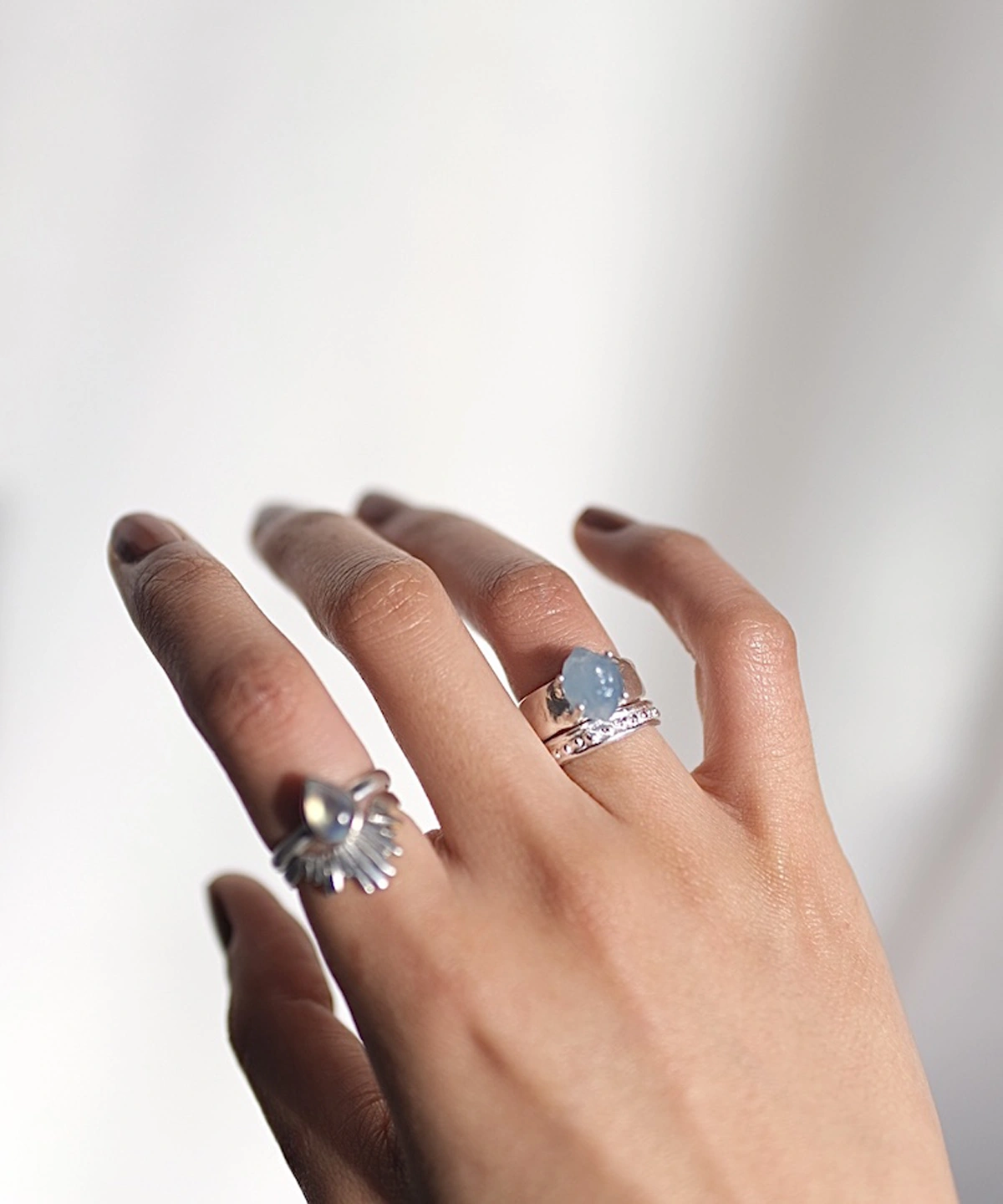 March Birthstone Ring-Natural Aquamarine Ring in Sterling Silver 03 Hand with brown nails wearing silver rings, including a March Birthstone Ring with oval natural aquamarine and a teardrop sunburst design, styled against a soft blurred background.