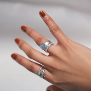 March Birthstone Ring-Natural Aquamarine Ring in Sterling Silver 04 Hand with brown nails wearing two sterling silver rings, including a March Birthstone Ring with raw natural aquamarine and teardrop blue stone, styled against soft white fabric.