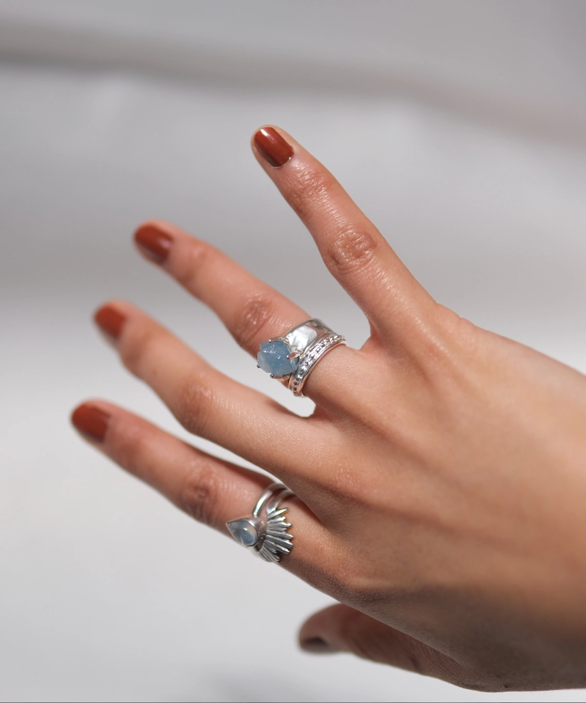 March Birthstone Ring-Natural Aquamarine Ring in Sterling Silver 04 Hand with brown nails wearing two sterling silver rings, including a March Birthstone Ring with raw natural aquamarine and teardrop blue stone, styled against soft white fabric.