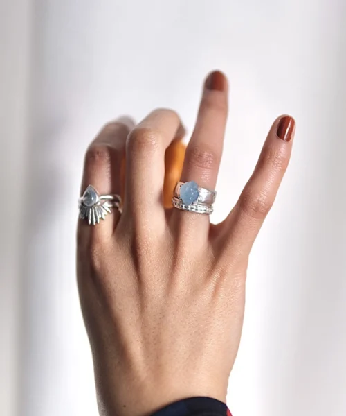 Hand with brown nails wearing silver rings, including a March Birthstone Ring with raw natural aquamarine, styled with sunburst and textured bands against a soft background.