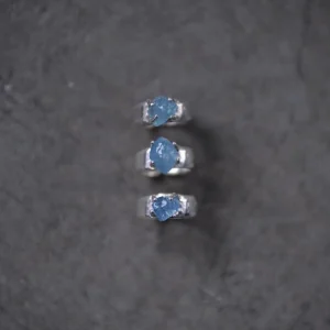 Three sterling silver rings with raw natural aquamarine gemstones arranged vertically on a dark textured surface, showcasing the March Birthstone Ring in organic prong settings.