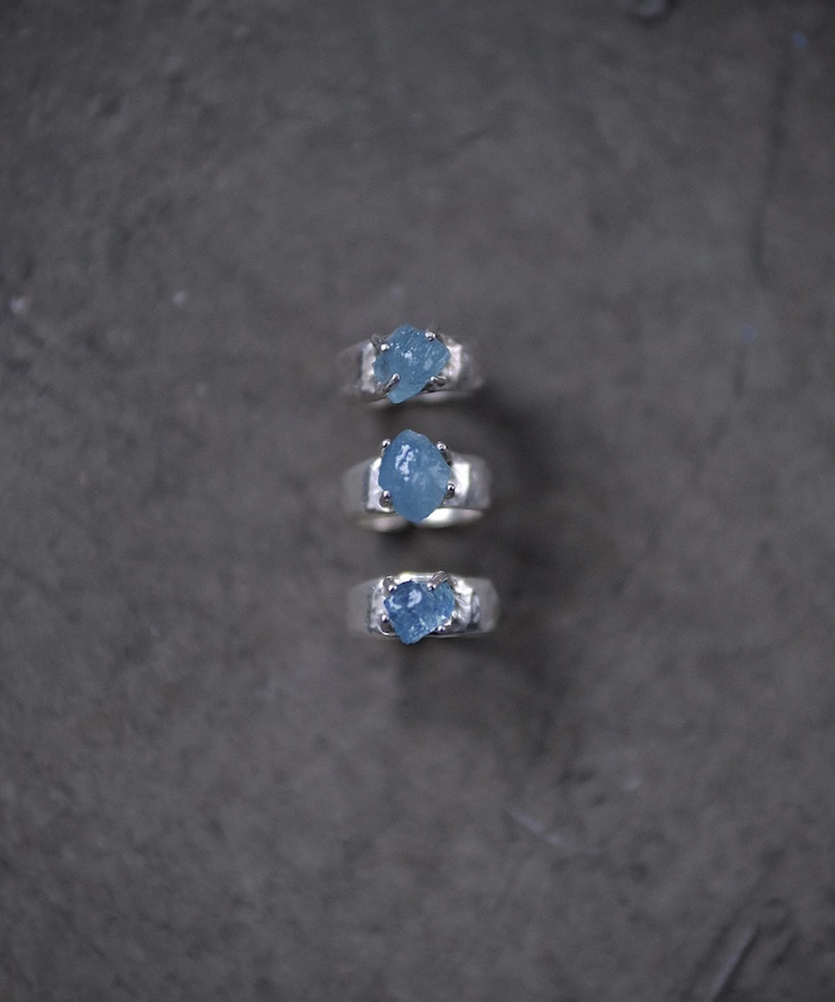 March Birthstone Ring-Natural Aquamarine Ring in Sterling Silver 07 Three sterling silver rings with raw natural aquamarine gemstones arranged vertically on a dark textured surface, showcasing the March Birthstone Ring in organic prong settings.