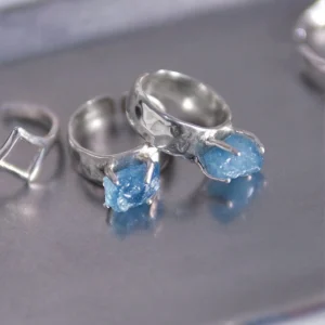 Three sterling silver rings on a metallic surface, including a March Birthstone Ring with raw natural aquamarine, styled with geometric and organic prong-set designs.