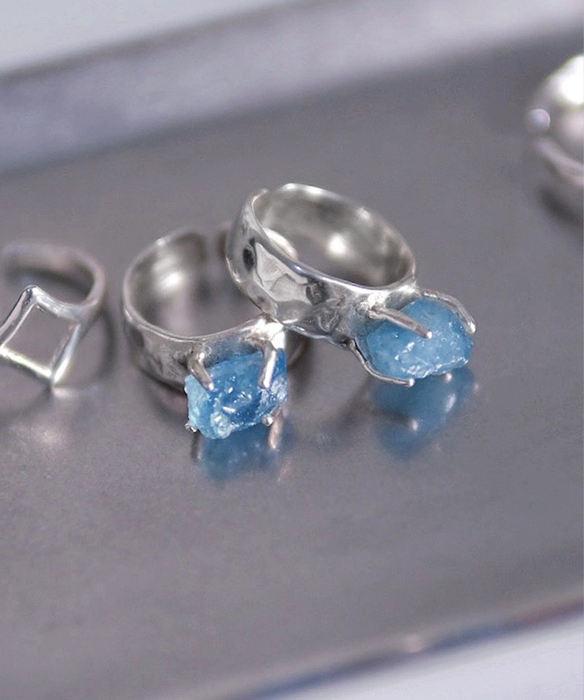 Three sterling silver rings on a metallic surface, including a March Birthstone Ring with raw natural aquamarine, styled with geometric and organic prong-set designs.