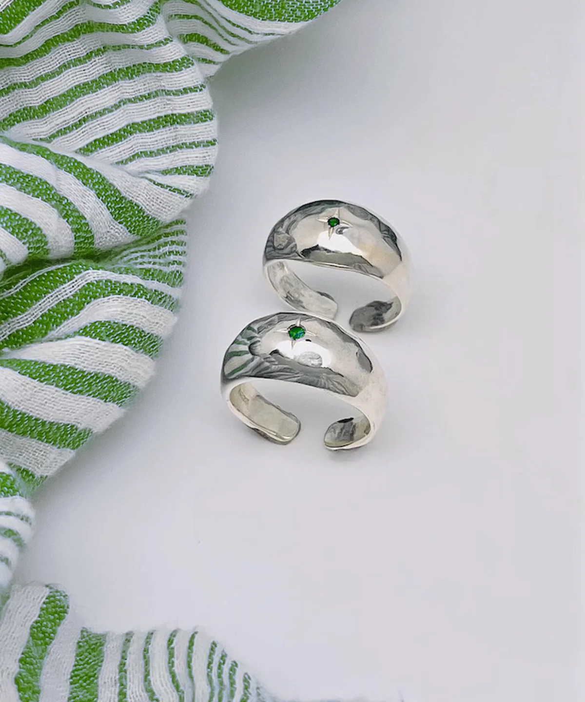 Two sterling silver open-ended rings with hammered texture and small natural emerald gemstones, placed on a white surface beside green-striped fabric, showcasing the May Birthstone Ring’s artisanal charm.