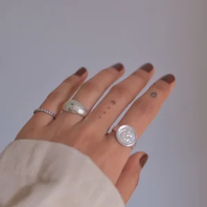 Hand with brown nails wearing silver rings, including a May Birthstone Ring with natural emerald and wax seal-style design, styled with minimalist tattoos and a soft sleeve.