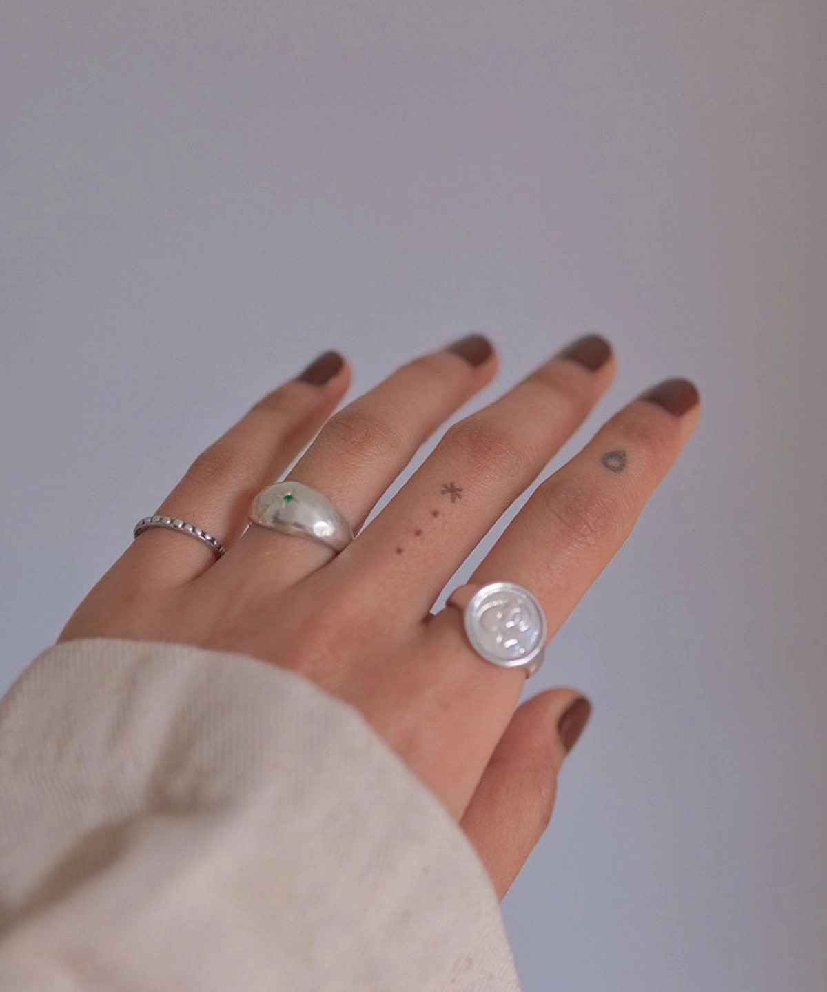 Hand with brown nails wearing silver rings, including a May Birthstone Ring with natural emerald and wax seal-style design, styled with minimalist tattoos and a soft sleeve.