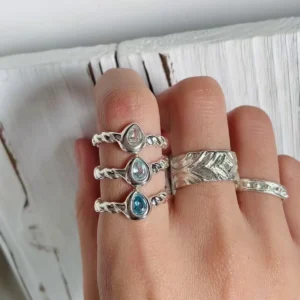 Hand wearing five sterling silver rings, featuring a November Birthstone Ring with a teardrop-shaped topaz, twisted bands, leaf engraving, and dotted textures, styled on a rustic whitewashed wood background.