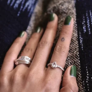 Hand with green nails wearing two sterling silver rings, including a twisted band November Birthstone Ring with topaz, set against a woven blue and beige fabric background.