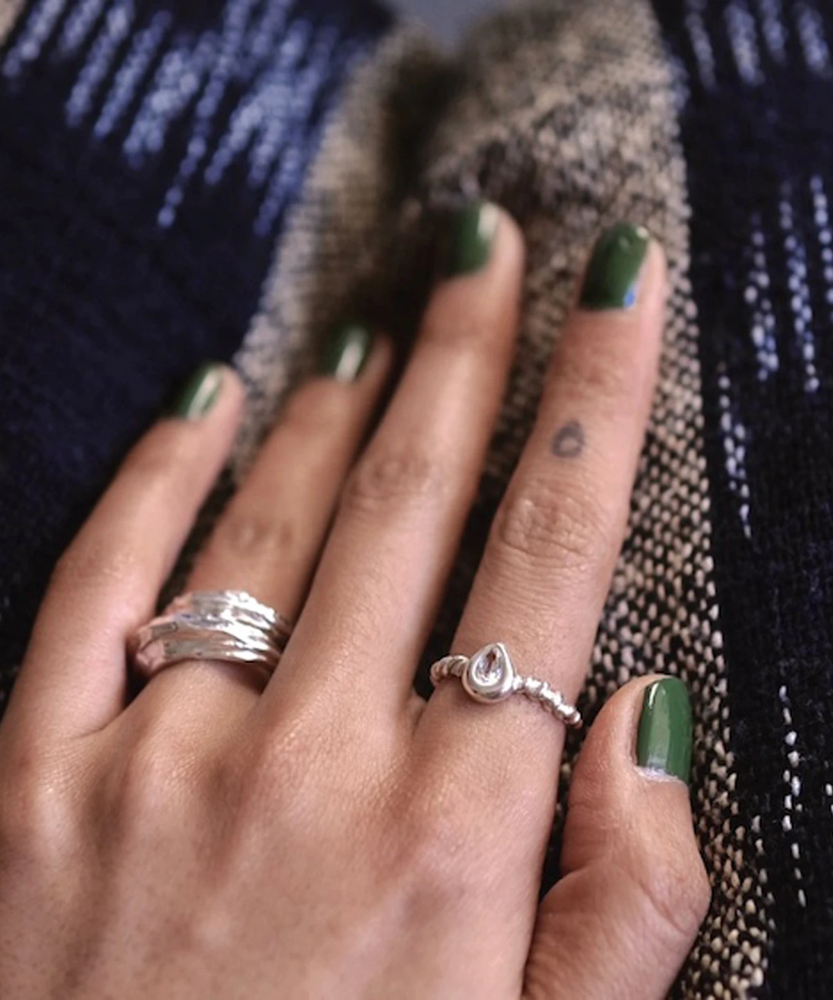 Hand with green nails wearing two sterling silver rings, including a twisted band November Birthstone Ring with topaz, set against a woven blue and beige fabric background.