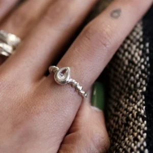 Hand with green-painted nails wearing a sterling silver November Birthstone Ring with topaz in a beaded band, set beside a teardrop tattoo and textured fabric background.