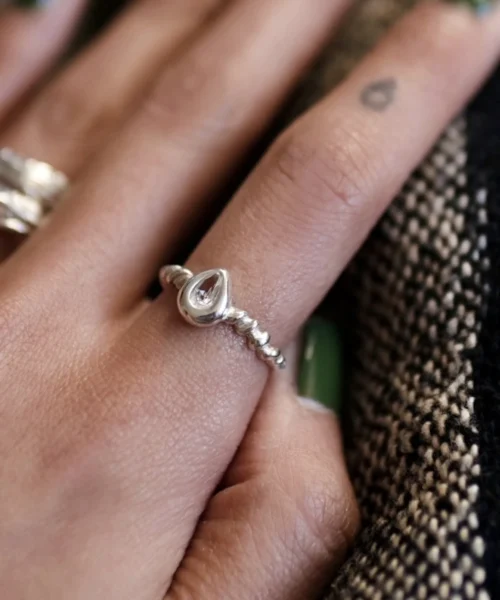 Hand with green-painted nails wearing a sterling silver November Birthstone Ring with topaz in a beaded band, set beside a teardrop tattoo and textured fabric background.