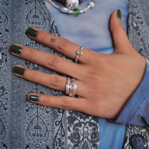Hand with dark green nails wearing sterling silver rings, including a November Birthstone Ring with topaz, styled with pearl accents, teardrop tattoo, and beaded necklace.