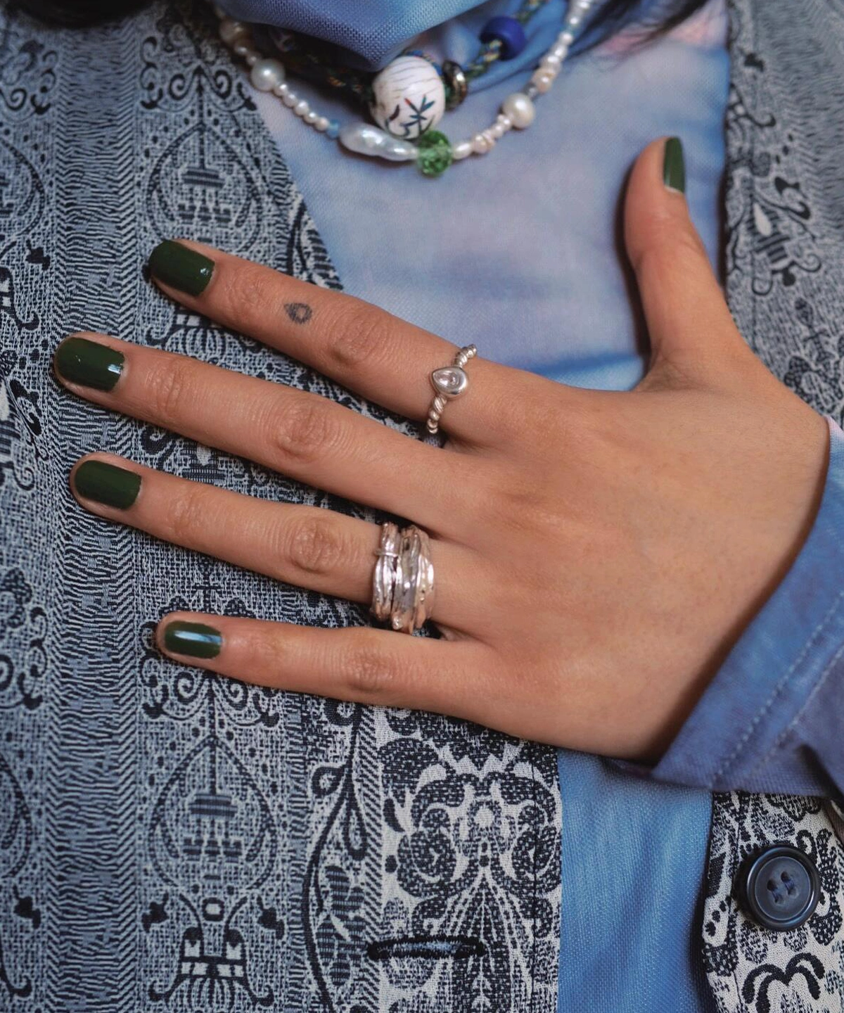 Hand with dark green nails wearing sterling silver rings, including a November Birthstone Ring with topaz, styled with pearl accents, teardrop tattoo, and beaded necklace.