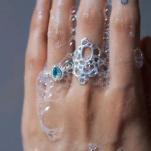 Hand covered in soap bubbles wearing sterling silver rings, including a November Birthstone Ring with topaz, styled with a blue teardrop tattoo and bubble-inspired design.