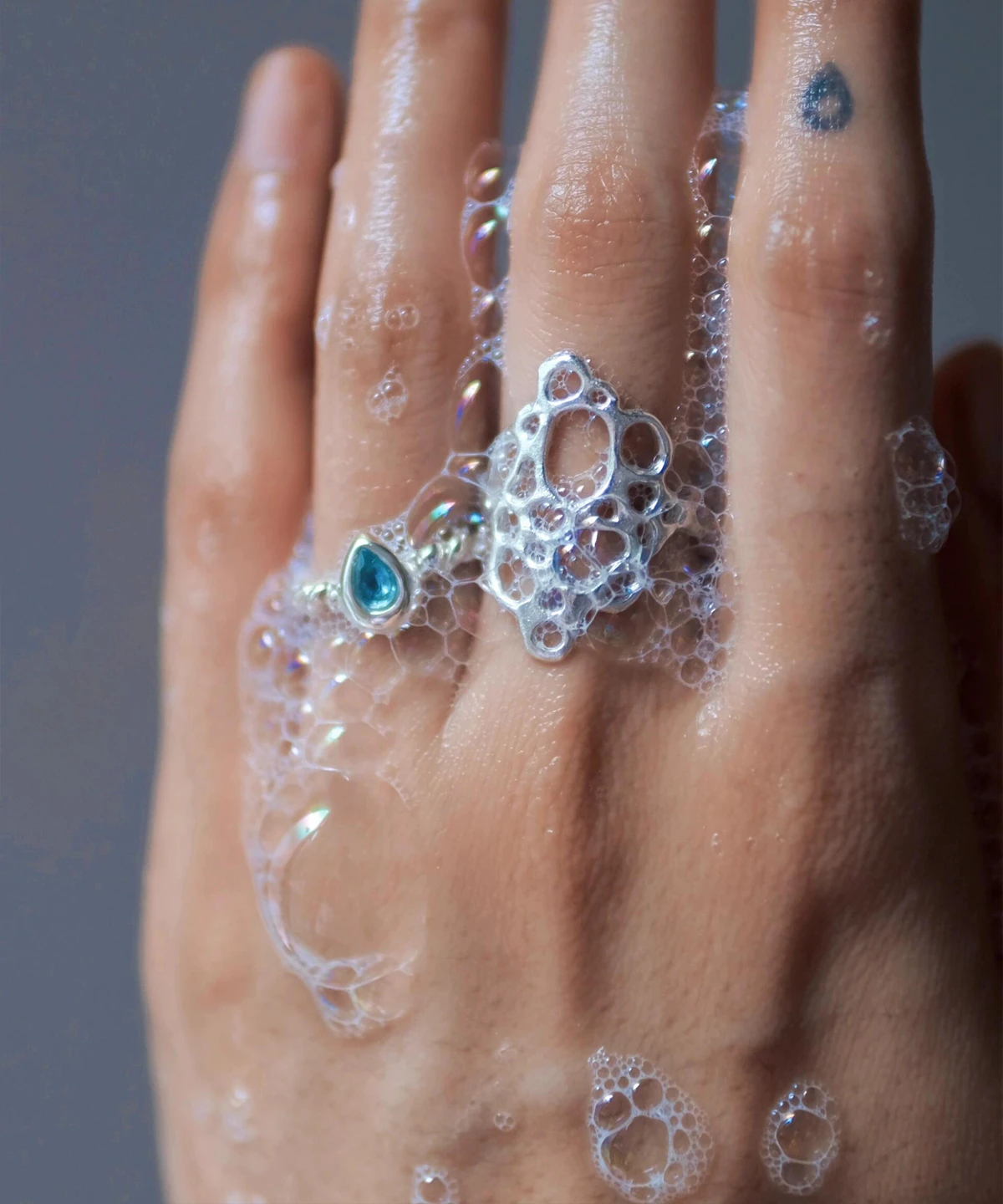 Hand covered in soap bubbles wearing sterling silver rings, including a November Birthstone Ring with topaz, styled with a blue teardrop tattoo and bubble-inspired design.