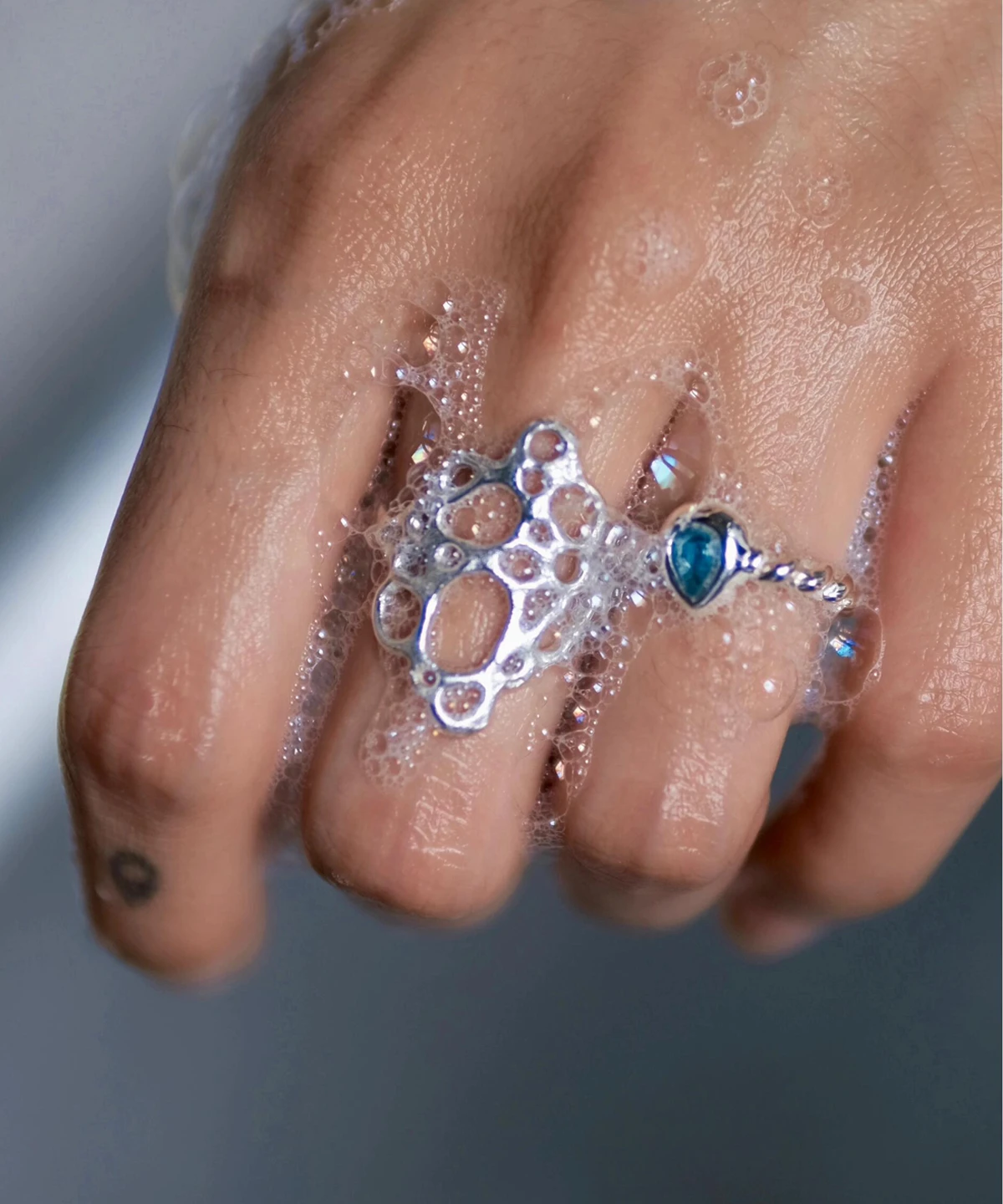 Hand covered in soap bubbles wearing sterling silver rings, including a November Birthstone Ring with topaz, styled with a circular tattoo and bubble-inspired textures.