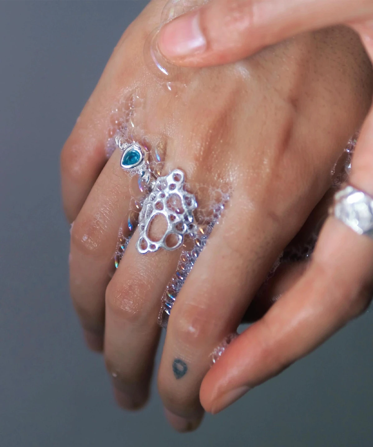 Hand covered in soap bubbles wearing sterling silver rings, including a November Birthstone Ring with topaz, styled with a blue gemstone and delicate tattoo for ethereal charm.
