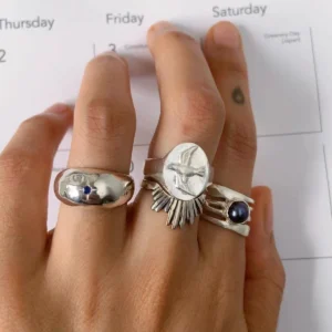 Hand with brown nails wearing silver rings, including a September Birthstone Ring with natural sapphire and bird engraving, styled over a calendar page with cultural references.