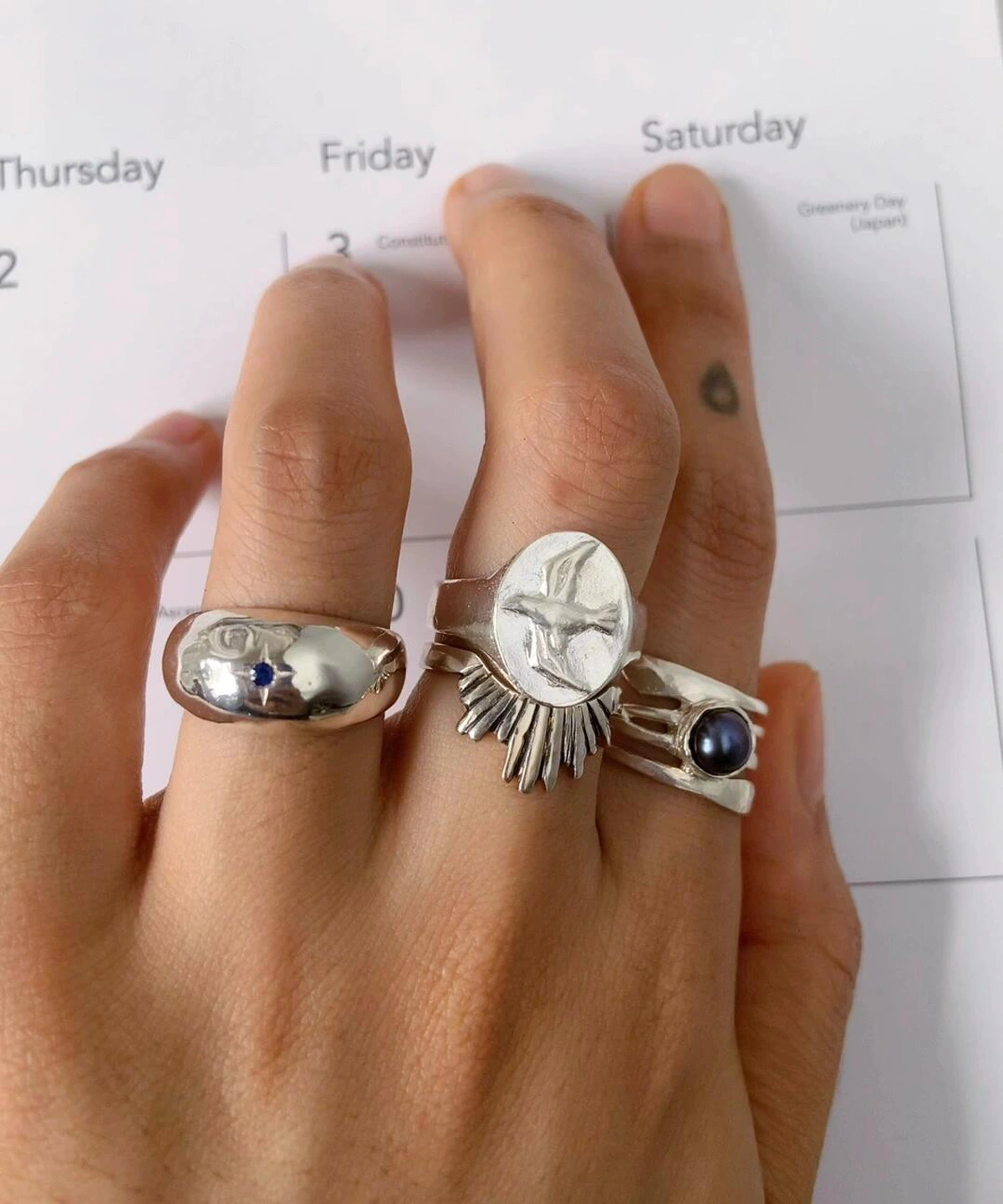 Hand with brown nails wearing silver rings, including a September Birthstone Ring with natural sapphire and bird engraving, styled over a calendar page with cultural references.