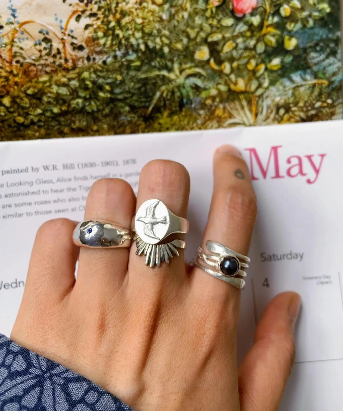 Hand with brown nails wearing silver rings, including a September Birthstone Ring with natural sapphire and snake motif, styled over a calendar page with floral art.