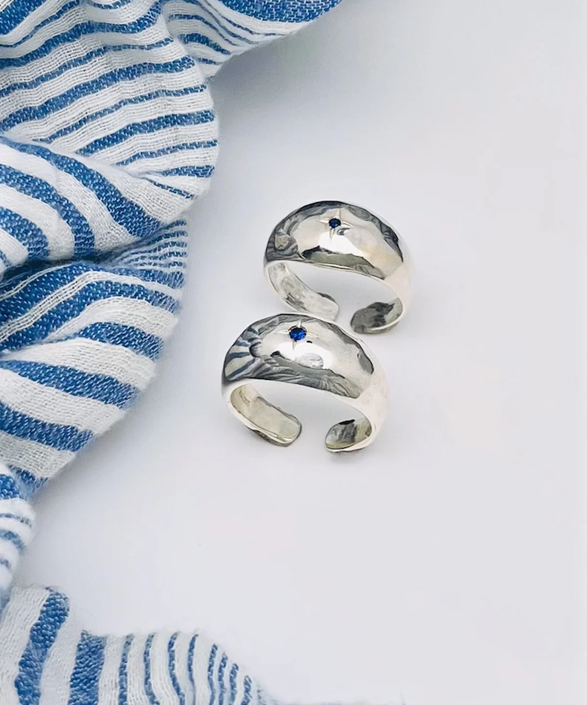 Two sterling silver open-ended rings with small natural sapphire gemstones, placed on a white surface beside striped fabric, showcasing the September Birthstone Ring’s artisanal charm.