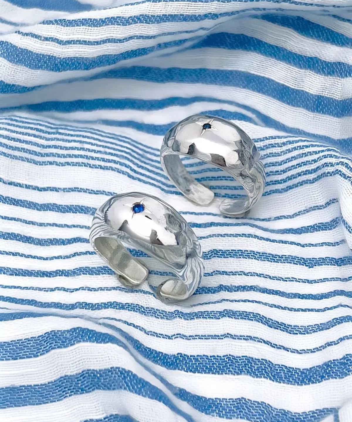 Two sterling silver open-ended rings with hammered texture and small natural sapphire gemstones, placed on striped fabric to showcase the September Birthstone Ring’s artisanal charm.