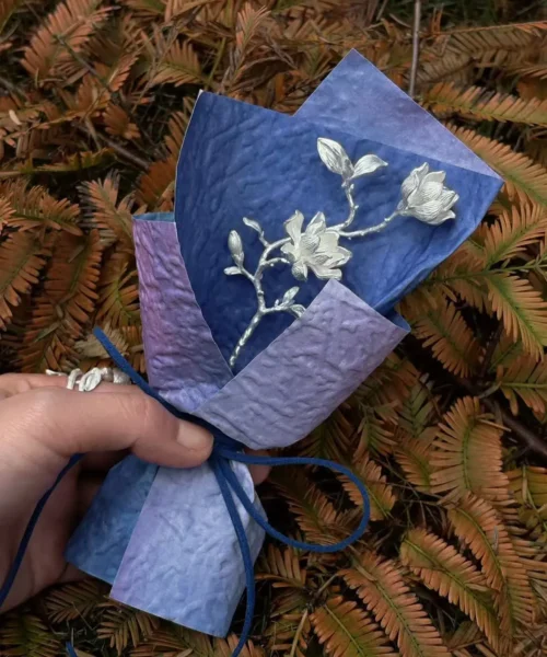 Hand holding a 925 Sterling Silver Magnolia Bouquet wrapped in purple and blue paper with dark blue string; a nature-inspired Handmade Art Gift featuring sculptural silver flowers and leaves, styled against fern foliage.