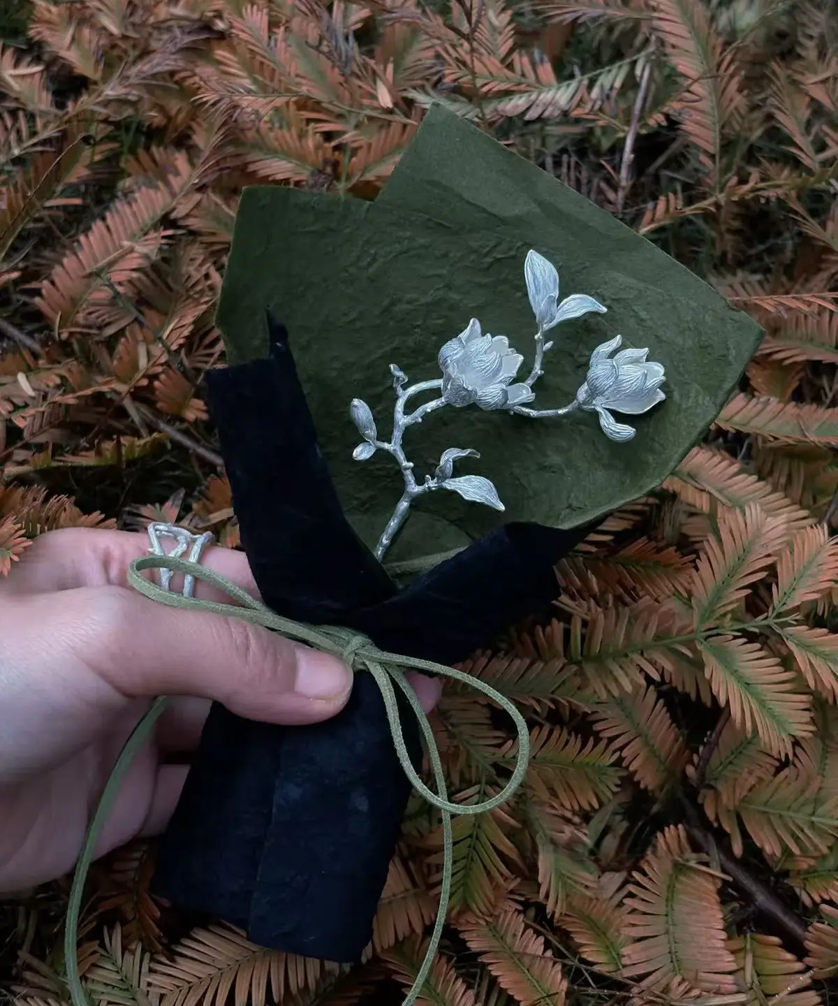 Hand holding a 925 Sterling Silver Magnolia Bouquet wrapped in black and green paper with green string; a nature-inspired Handmade Art Gift featuring sculptural silver magnolia blossoms and leaves, styled against fern foliage.