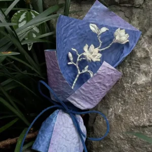 925 Sterling Silver Magnolia Bouquet wrapped in gradient blue and purple paper with silver floral branch, tied with blue ribbon; a nature-inspired Handmade Art Gift styled against rocks and green foliage.